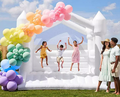 A white inflatable bounce house with a sleek, modern design, featuring a front entrance, safety netting, and a small attached slide. The bounce house is set up outdoors on a flat surface, ideal for children's parties, weddings, and upscale events.